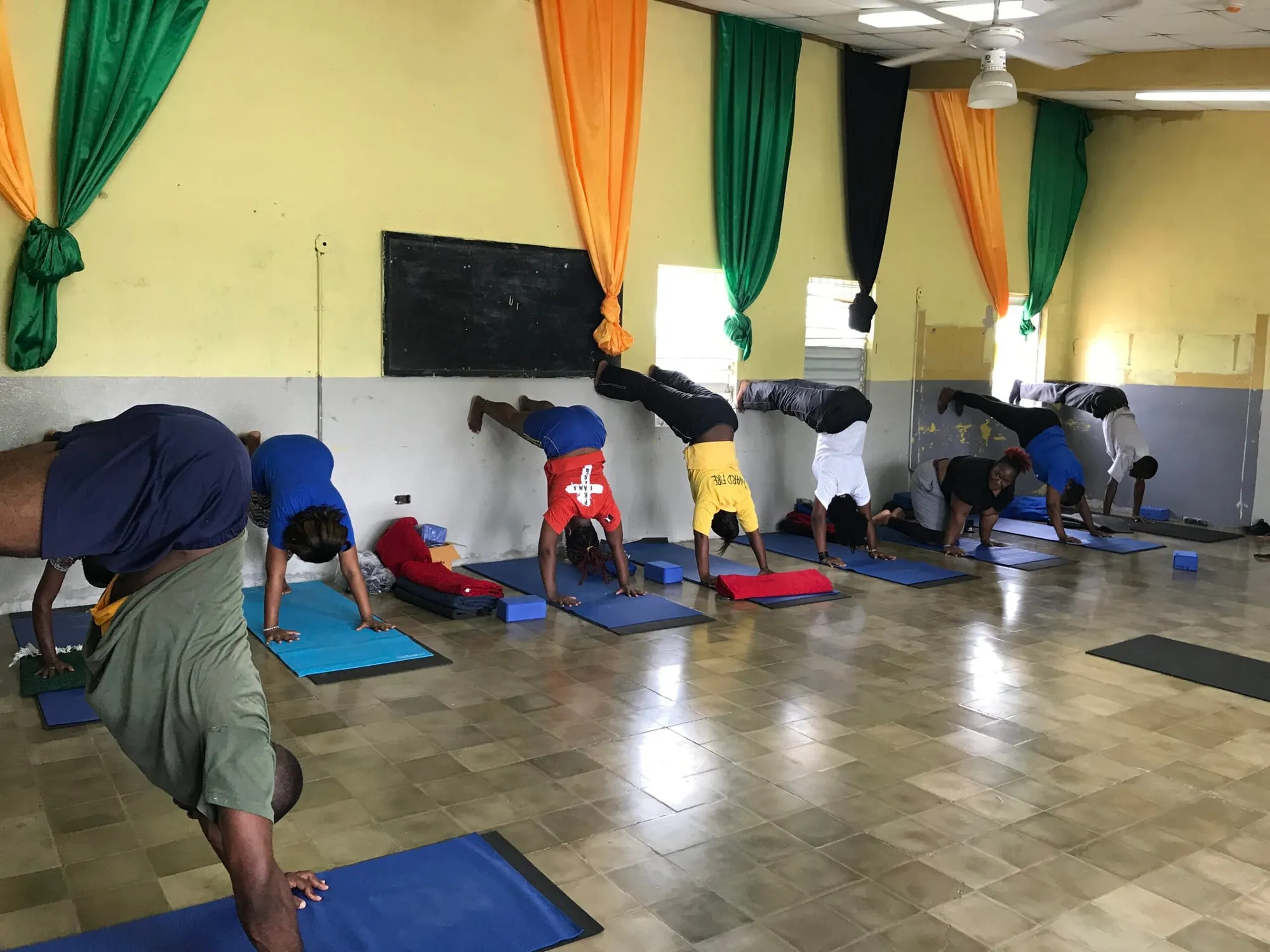 A group holding a supported pose on blue mats in a classroom hung with green, gold and black curtains