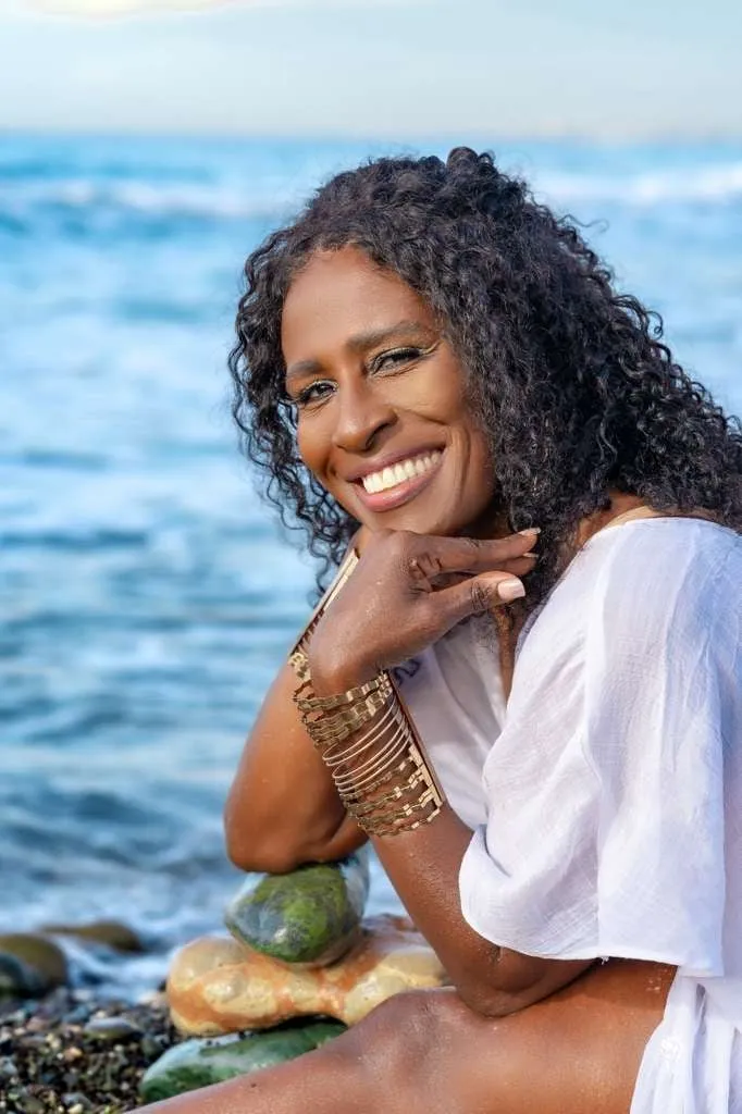 Sandra “Subhadra” Griffiths smiling, seated on stones by the sea